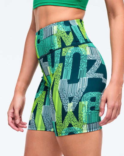 High-waisted biker shorts with a wide waistband and allover teal, lime, and navy graphic print, shown in a close side view on a female model wearing a green sports bra on a white studio background.