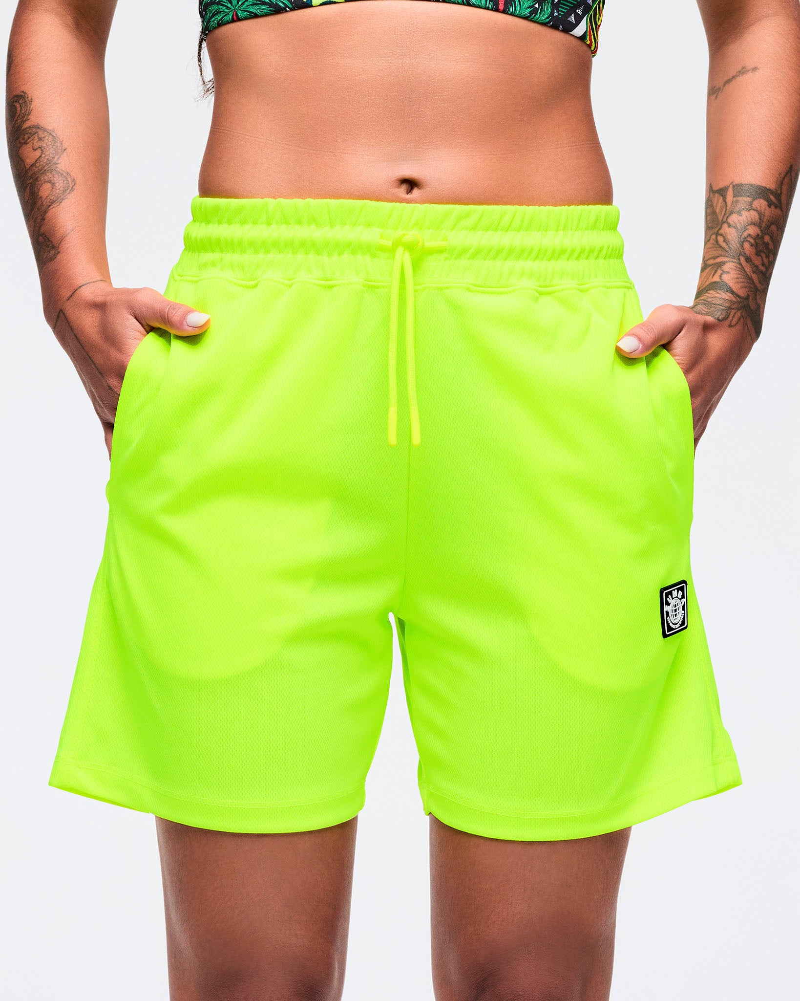 Neon lime mesh athletic shorts with an elastic waistband and adjustable drawcord, shown in a close-up front view on a female model with hands in pockets against a white studio background.