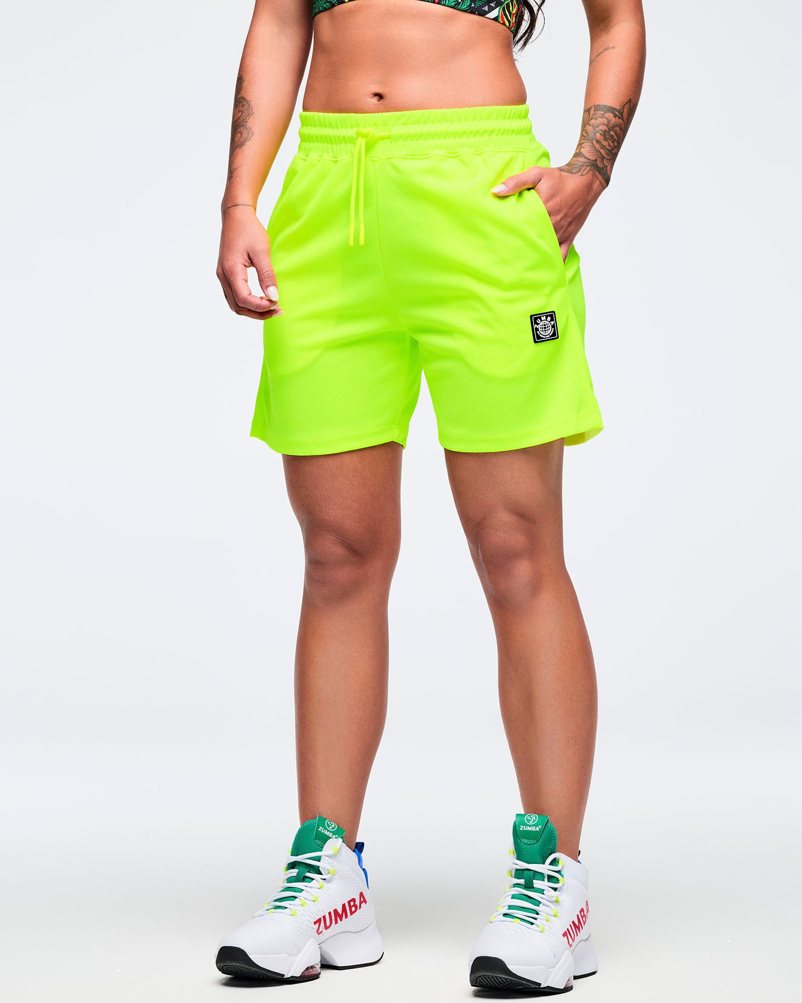 Neon lime mesh athletic shorts with elastic drawstring waistband, side pockets, and a small Zumba logo patch near the hem, modeled by a female wearing white high-top athletic sneakers on a white studio background.