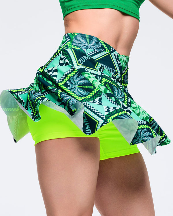 Green Zumba printed skort with flared hem and built-in neon lime shorts, captured in motion to show the swing of the skirt. Female model wears a solid green sports bra on a light gray studio background.