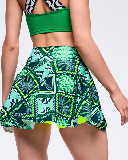 Green Zumba printed skort with flared hem and allover patchwork pattern in teal, navy, and lime, shown from the back. Female model wears it with a solid green sports bra on a light gray studio background.