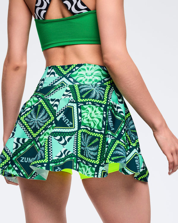 Green Zumba printed skort with flared hem and allover patchwork pattern in teal, navy, and lime, shown from the back. Female model wears it with a solid green sports bra on a light gray studio background.