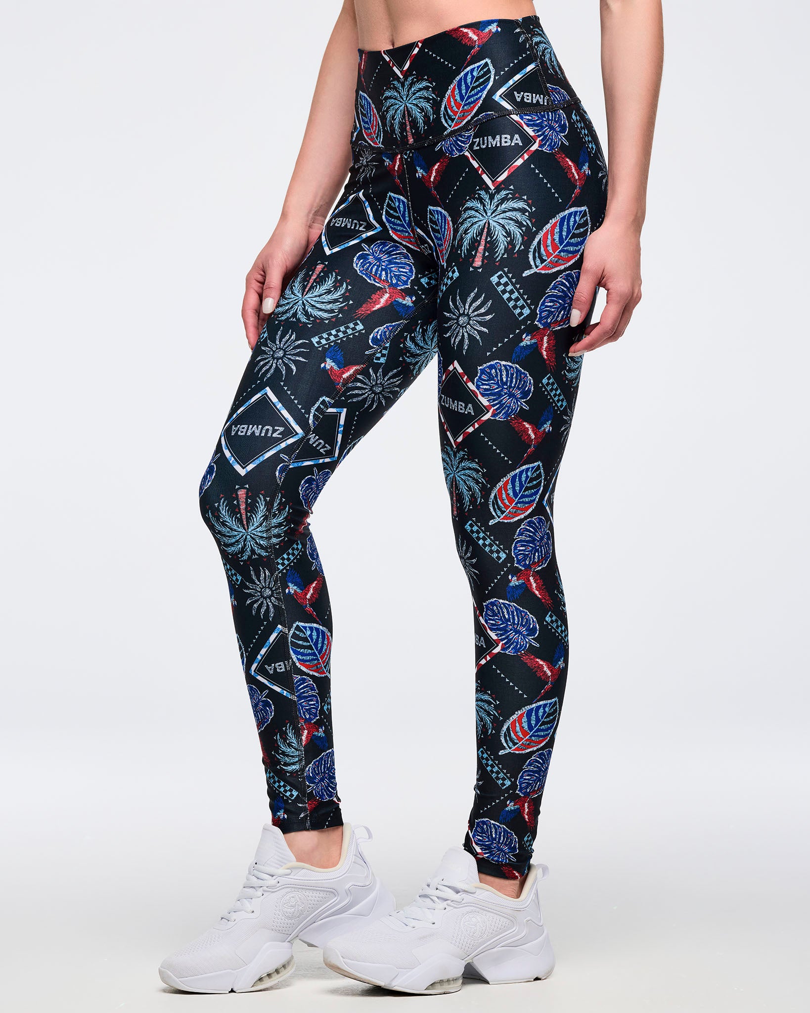 High-waisted black leggings with allover tropical print featuring palms, leaves, parrots, and “ZUMBA” diamond logos in red, blue, and teal; female model wears white sneakers, shown in a three-quarter front view on a white background.