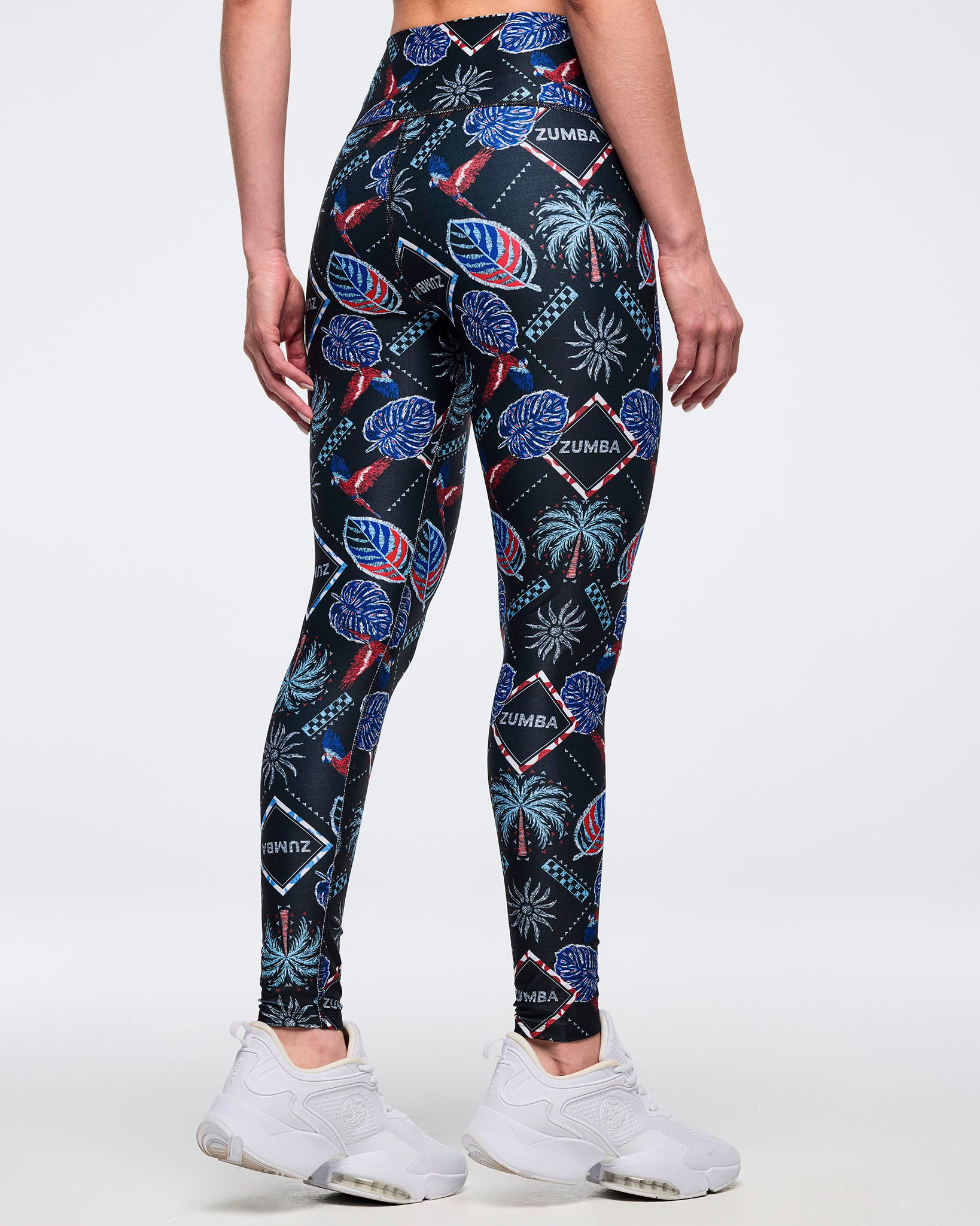 Back view of high-waisted black tropical-print leggings with “ZUMBA” diamond logos and palm-and-parrot graphics; female model wears white sneakers against a white background.