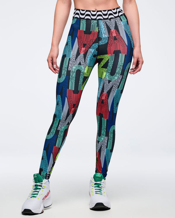 Front view of the multicolor letter-print leggings featuring oversized letter graphics and a black-and-white wavy waistband; female model wears white high-top Zumba sneakers on a white background.