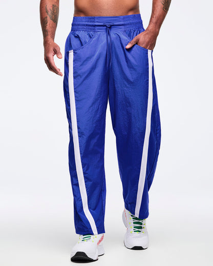 Front view of royal blue track pants with white side stripes, elastic drawstring waistband, and front pockets; male model wears white high-top Zumba sneakers on a white background.