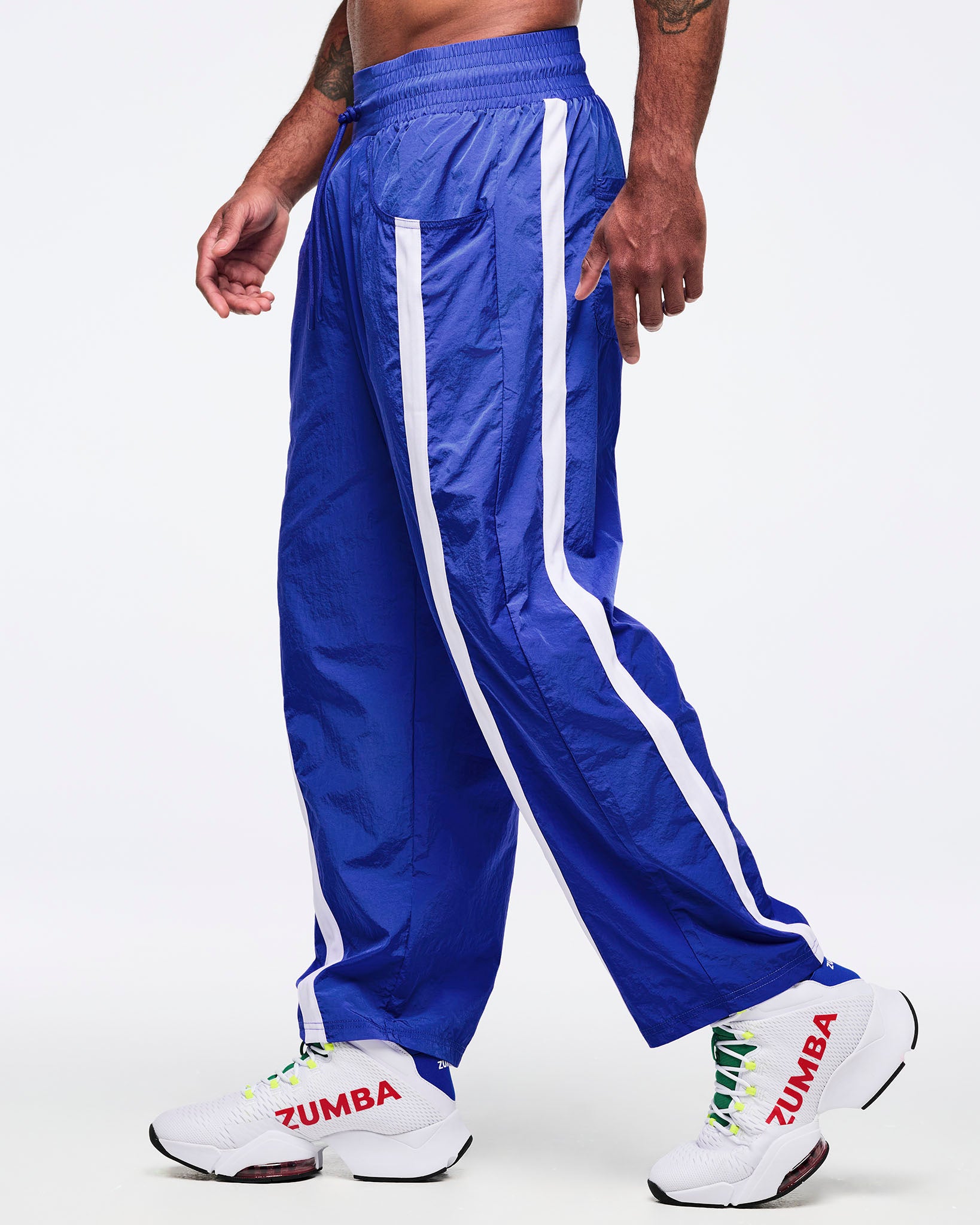 Side view of royal blue track pants with white side stripes and front pocket detail; male model wears white high-top Zumba sneakers, shown in motion on a white background.
