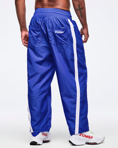 Back view of royal blue track pants with white side stripes and back patch pockets labeled “ZUMBA”; male model wears white sneakers on a white background.
