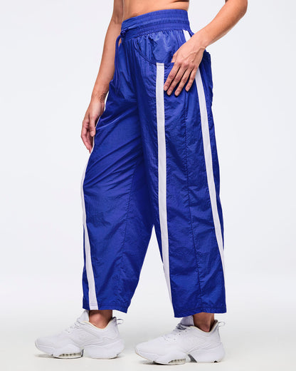 Royal blue nylon wide-leg track pants with white side stripes, elastic drawstring waistband, and front patch pockets; female model wears white sneakers, shown on a white background.
