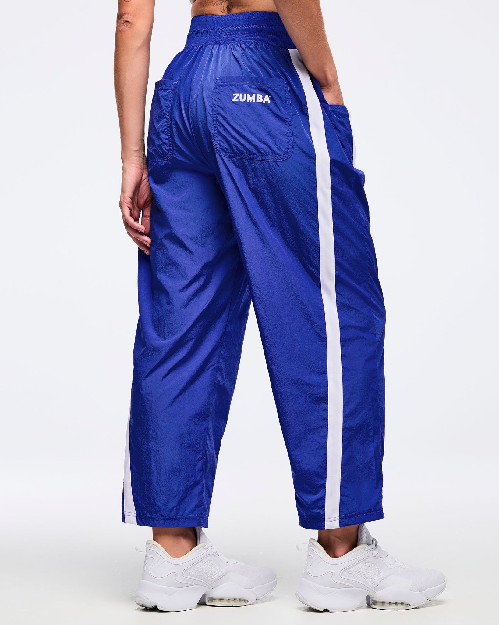 Back view of royal blue track pants with white side stripes, elastic waistband, and back patch pockets with “ZUMBA” text; female model wears white sneakers on a white background.
