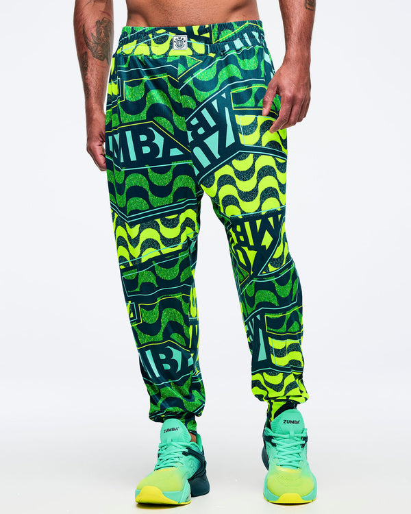 Front view of a male model wearing the neon green/teal graphic Zumba jogger pants; teal-and-lime Zumba sneakers on a white background.