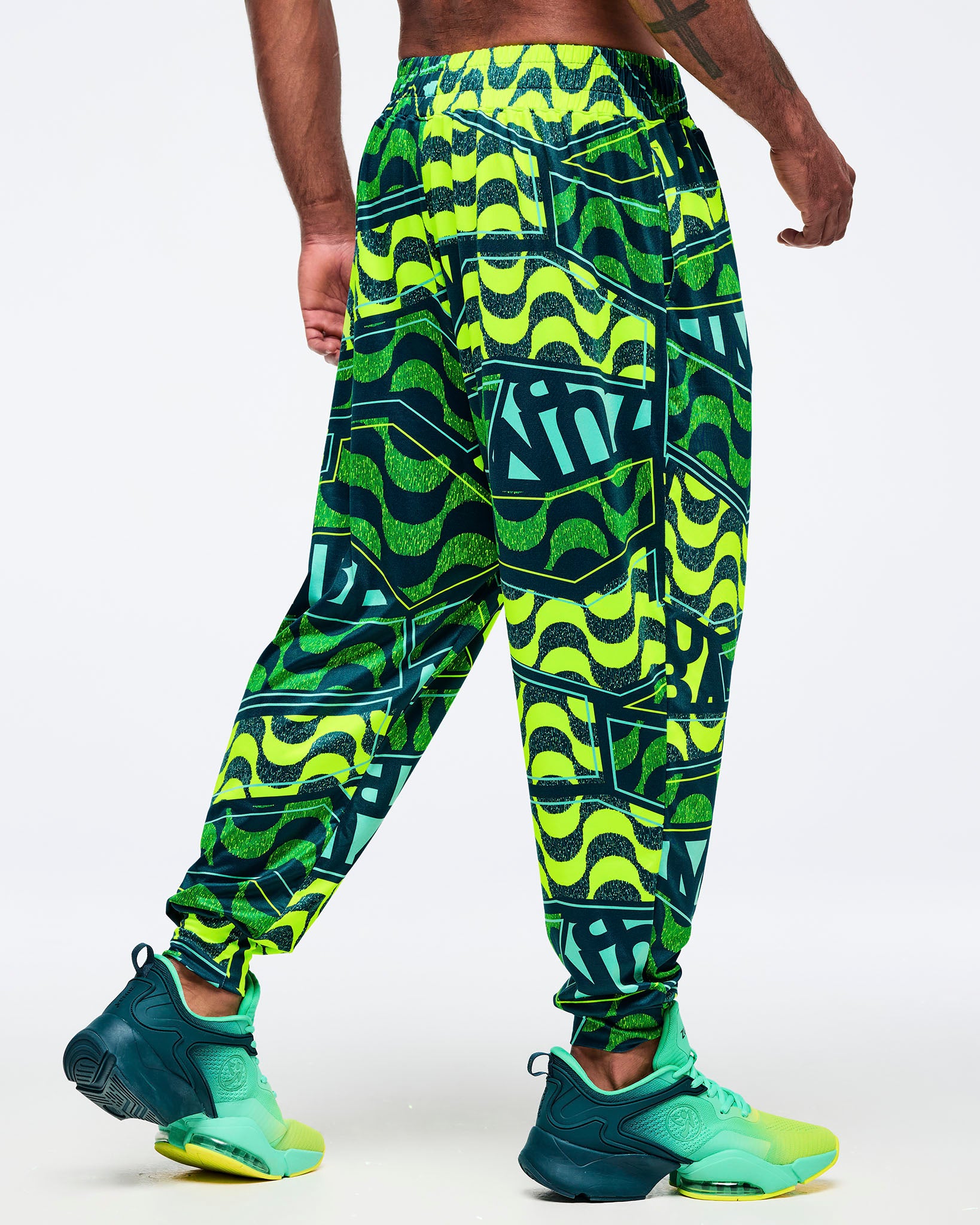 View of a male model wearing the neon green/teal graphic Zumba jogger pants; teal-and-lime Zumba sneakers on a white background.