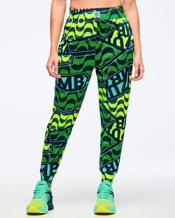 Straight-on front view of the neon green/teal/dark green graphic Zumba jogger pants (all-over wavy pattern + bold “ZUMBA” lettering), elastic waistband, tapered cuffs; teal-and-lime Zumba sneakers on a white background.