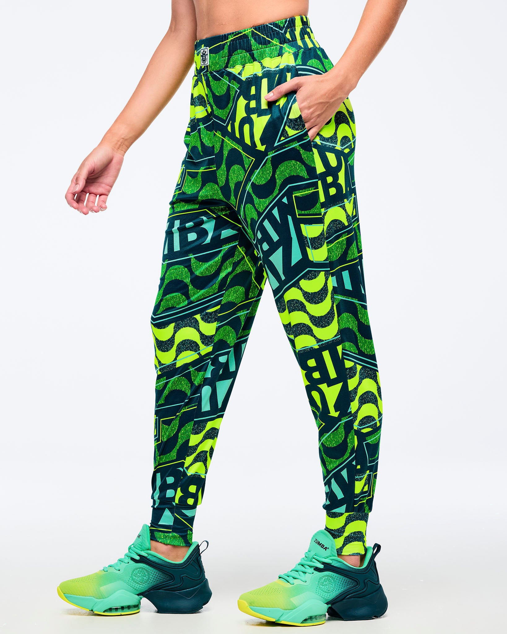 Front view of neon green, teal, and dark green Zumba jogger-style pants with an all-over wavy/graphic “ZUMBA” print and elastic waistband; paired with teal-and-lime Zumba sneakers on a white background.