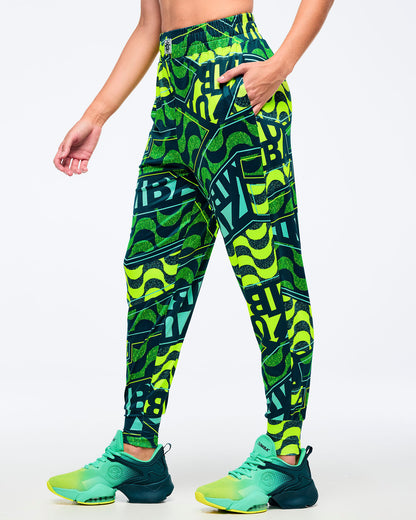 Front view of neon green, teal, and dark green Zumba jogger-style pants with an all-over wavy/graphic “ZUMBA” print and elastic waistband; paired with teal-and-lime Zumba sneakers on a white background.