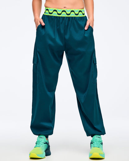 Teal cargo jogger pants with a neon green-and-black wave print elastic waistband, shown from the front on a female model with hands in pockets; loose legs with cargo pockets and elastic cuffs, paired with aqua-to-lime sneakers.