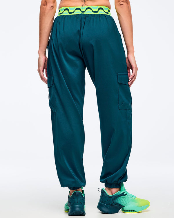 Teal cargo jogger pants with a neon green-and-black wave print elastic waistband, shown from the back on a female model; relaxed fit with gathered cuffs, side cargo pockets, and aqua-to-lime sneakers.