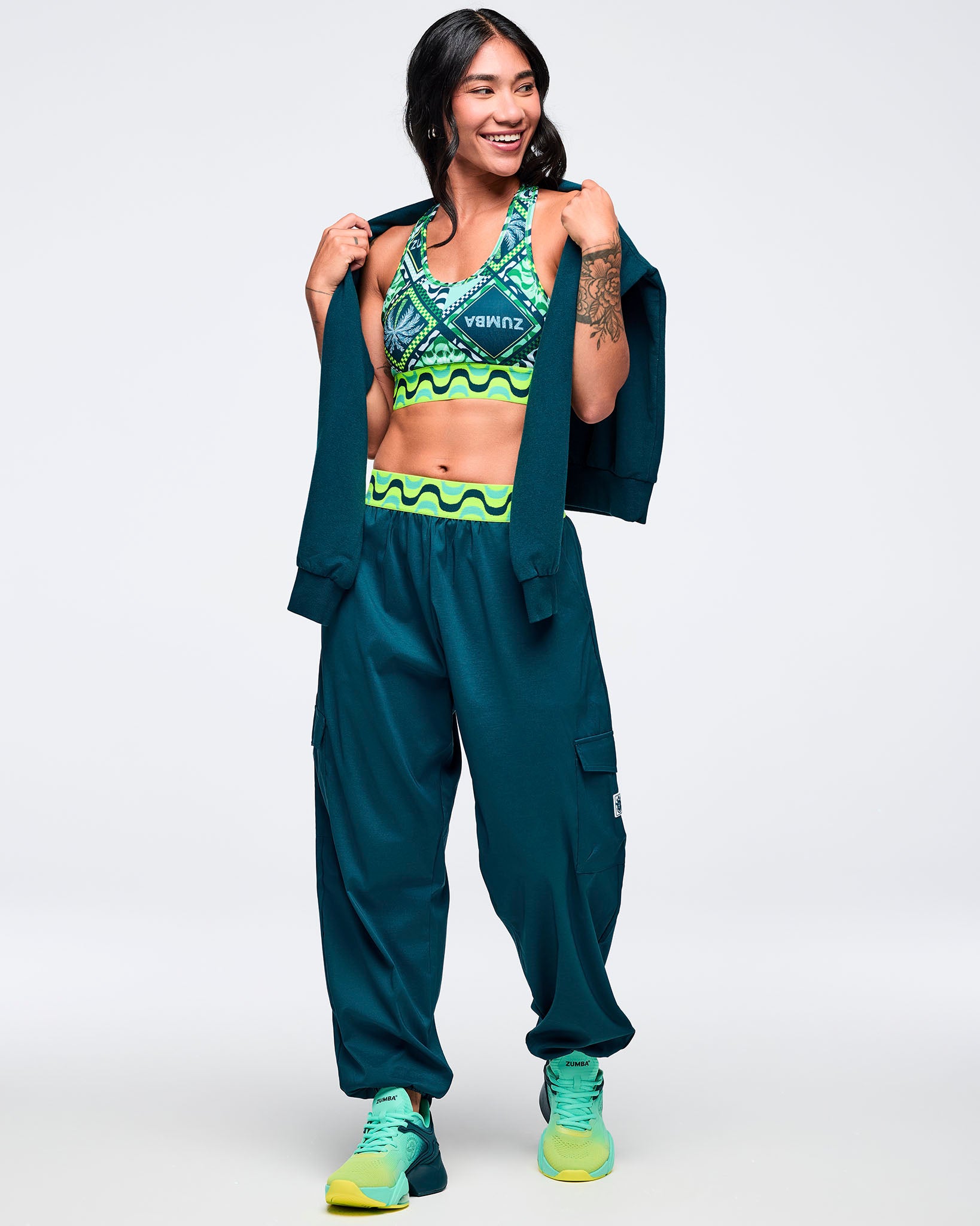 Teal cargo jogger pants with a neon wave print waistband, worn by a female model with a matching green patterned sports bra and teal jacket draped over her shoulders; paired with aqua-to-lime sneakers.