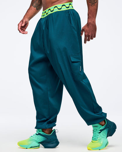 Teal cargo jogger pants with a neon green-and-black wave print waistband, shown on a male model in side view; relaxed fit with thigh cargo pocket and elastic cuffs, paired with aqua-to-lime sneakers.