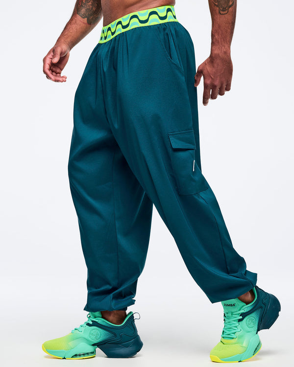 Teal cargo jogger pants with a neon green-and-black wave print waistband, shown on a male model in side view; relaxed fit with thigh cargo pocket and elastic cuffs, paired with aqua-to-lime sneakers.