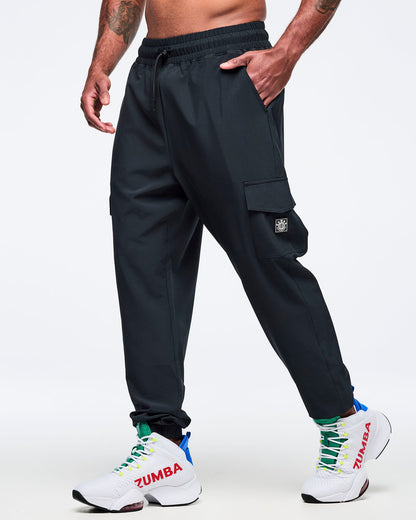 Black cargo jogger pants with an elastic drawstring waistband, worn by a male model in side view with one hand in a pocket; cargo pocket and ankle cuffs, paired with white high-top Zumba sneakers.