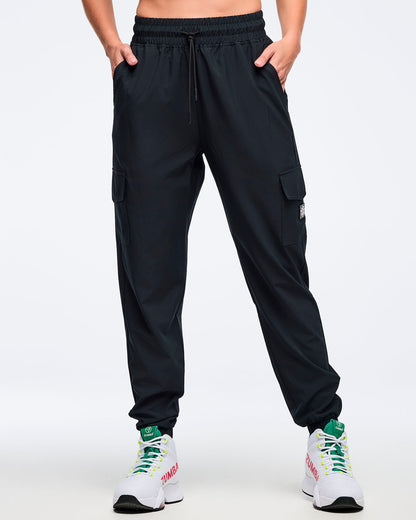 Black cargo jogger pants with an elastic drawstring waistband, shown from the front on a female model with hands in pockets; straight-to-tapered legs with cargo pockets and ankle cuffs, paired with white high-top Zumba sneakers.