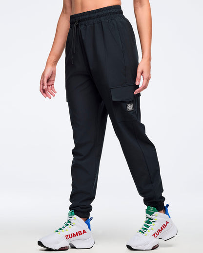 Black cargo jogger pants with an elastic drawstring waistband and tapered legs, shown on a female model in a side pose; flap cargo pocket and small logo patch on the thigh, paired with white high-top Zumba sneakers.
