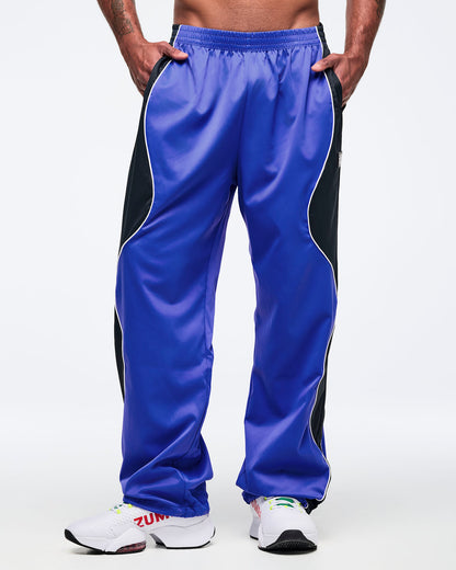 Blue and black track pants with white piping, elastic waistband, and roomy straight-leg cut, shown from the front on a male model with hands in pockets and white athletic sneakers.