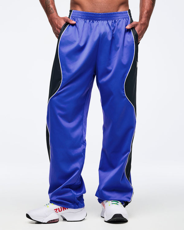 Blue and black track pants with white piping, elastic waistband, and roomy straight-leg cut, shown from the front on a male model with hands in pockets and white athletic sneakers.