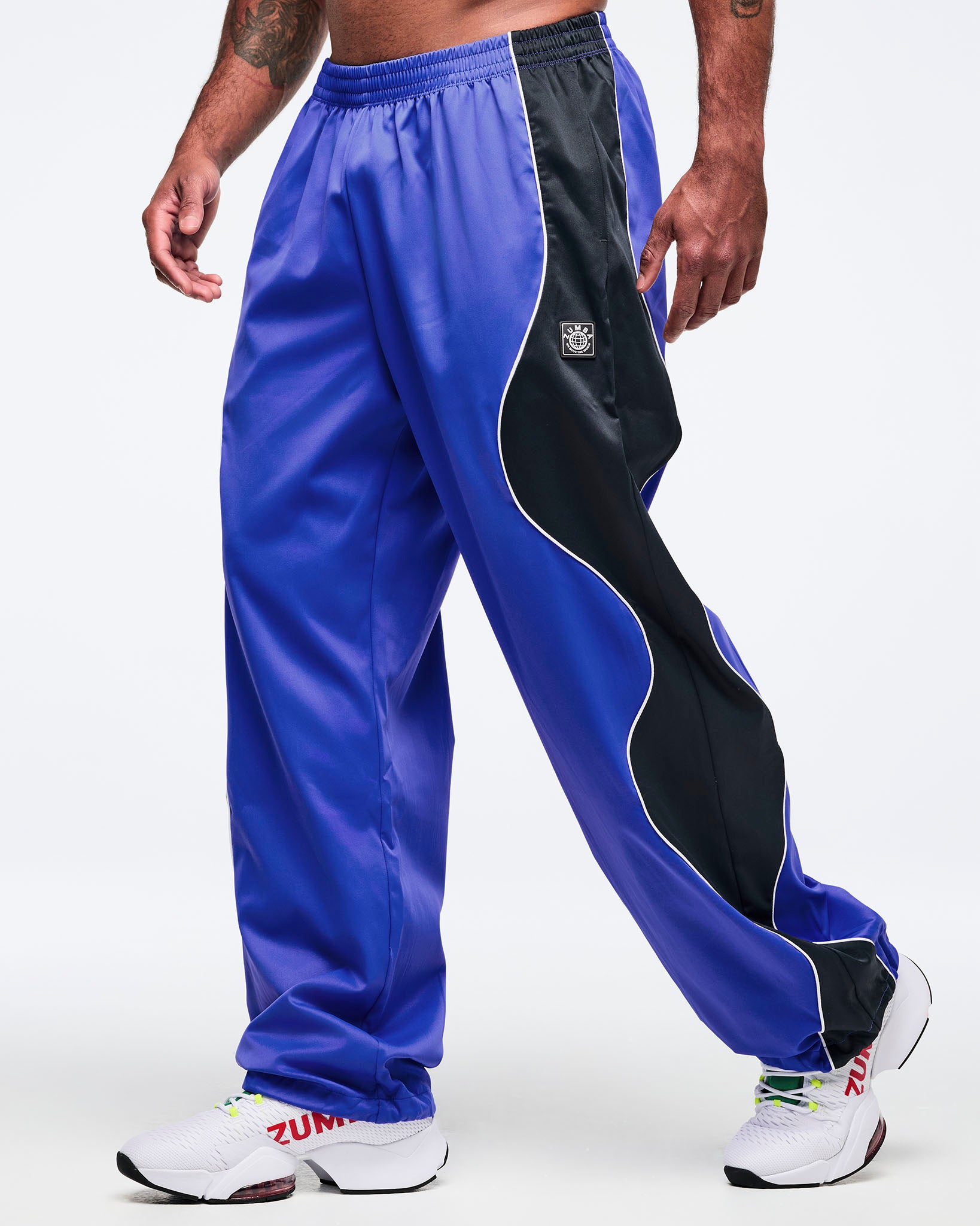 Blue track pants with black side paneling, white piping, and elastic waistband in a loose straight-leg cut, shown at an angle on a male model wearing white athletic sneakers.