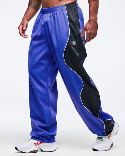 Blue track pants with black side paneling, white piping, and elastic waistband in a loose straight-leg cut, shown at an angle on a male model wearing white athletic sneakers.