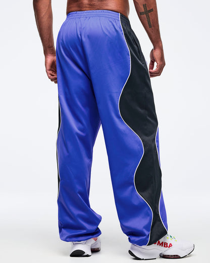 Blue track pants with black side panel and white piping, elastic waistband, and loose straight-leg fit, shown from the back on a male model wearing white athletic sneakers.
