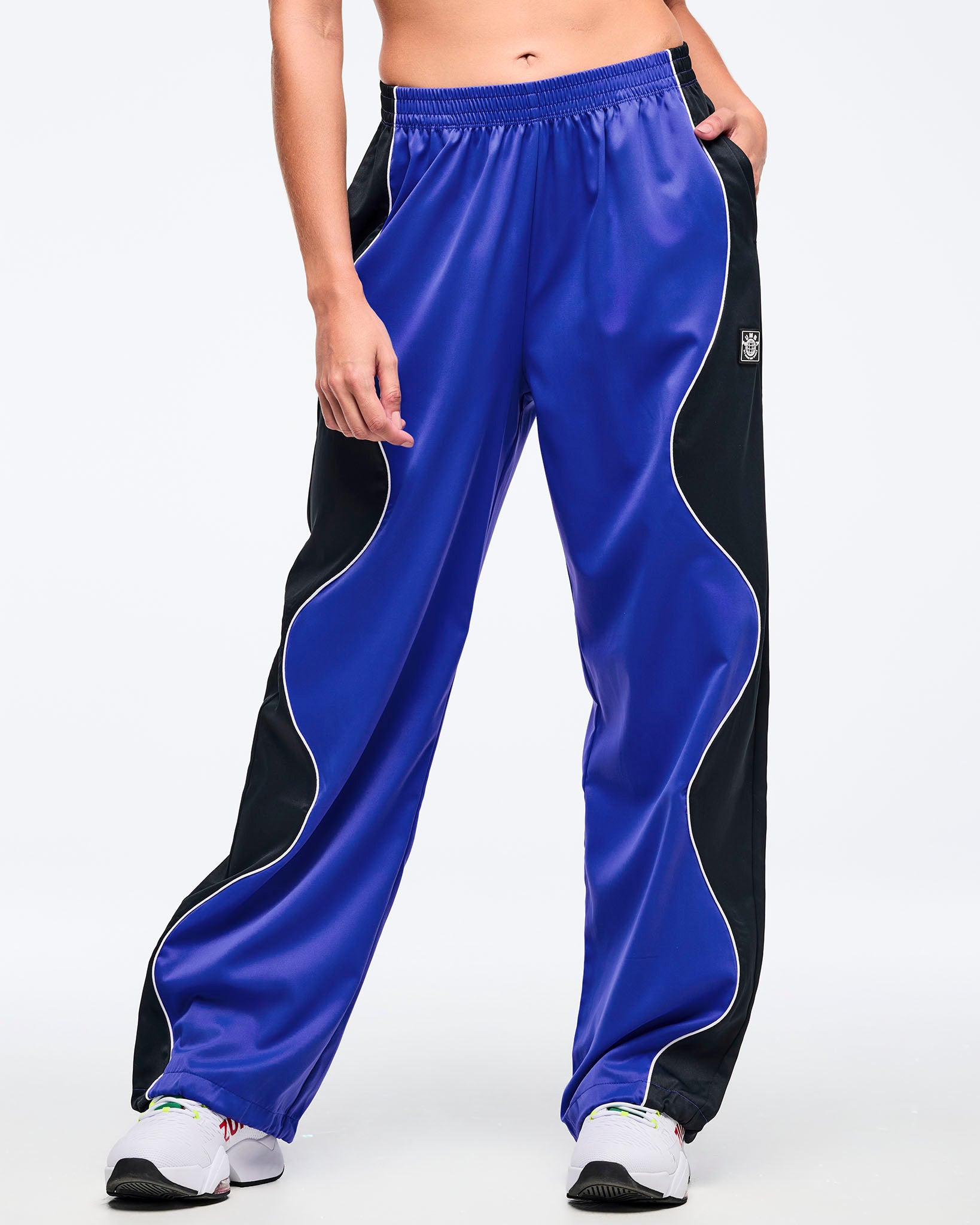 Blue track pants with black side panels, white piping, elastic waistband, and relaxed fit, shown from the front on a female model wearing white athletic sneakers with one hand in the pocket.