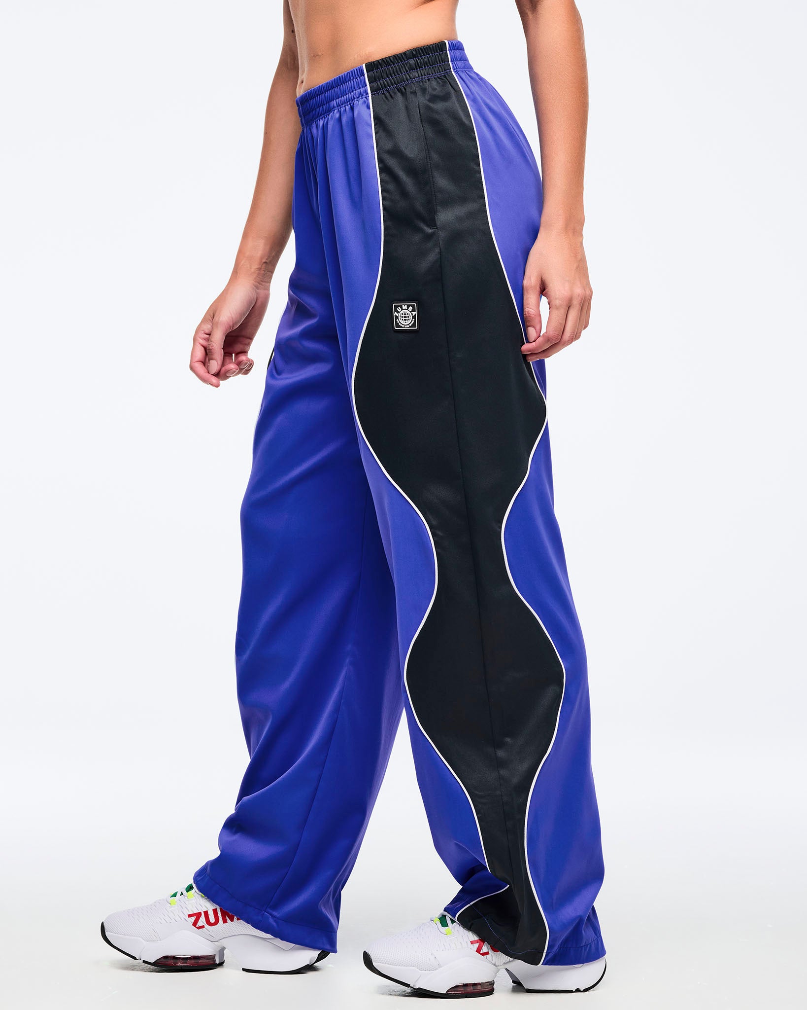 Blue and black track pants with white piping, elastic waistband, and wide straight-leg fit, shown from the side on a female model wearing white athletic sneakers.