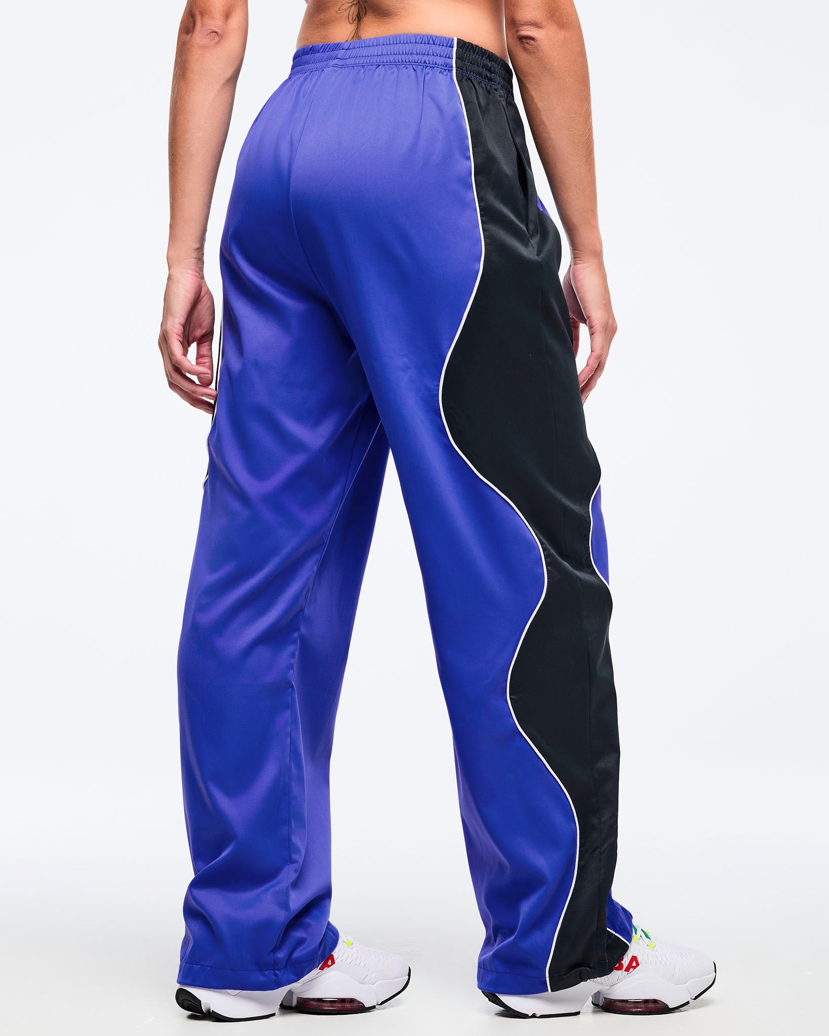 Blue track pants with a black side panel and white piping, elastic waistband, and relaxed straight-leg fit, shown from the back on a female model wearing white athletic sneakers.