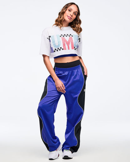 Blue and black track pants with white piping and elastic waistband, styled on a female model wearing a white cropped graphic T-shirt and white athletic sneakers, standing on a white studio background.