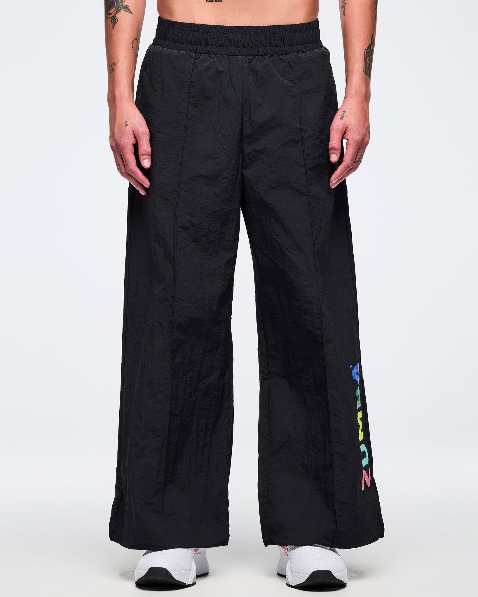 Front view of black wide-leg Zumba pants with elastic waistband and wide-leg cut; male model wears white sneakers on a white background.