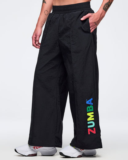 Black wide-leg Zumba pants with elastic waistband and multicolor vertical “ZUMBA” logo near the hem; male model wears white sneakers on a white background.