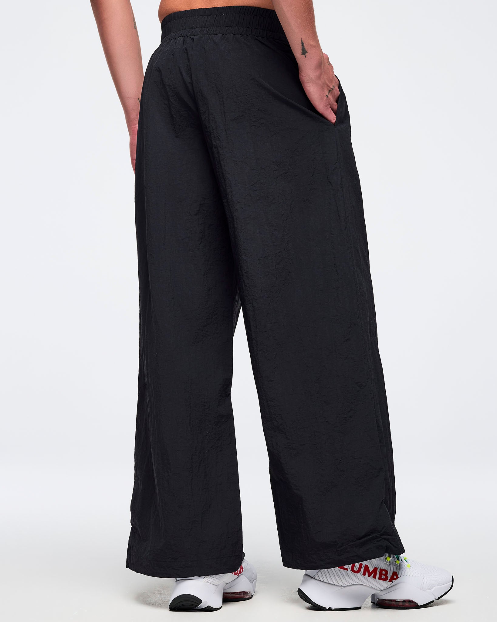 Back view of black wide-leg Zumba pants with elastic waistband and side pockets; male model wears white sneakers on a white background.