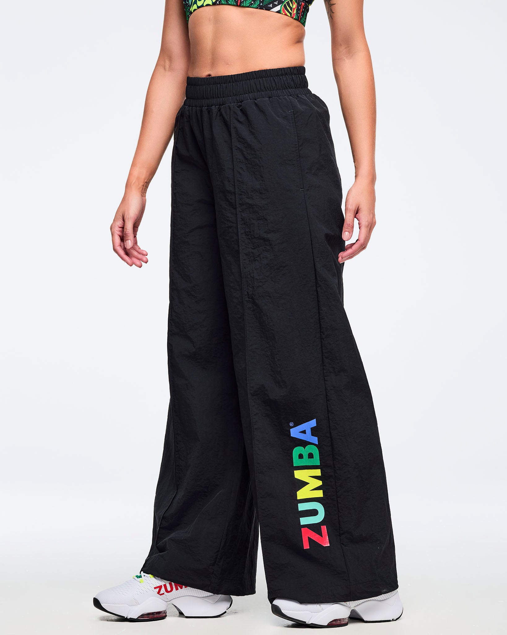 Black wide-leg Zumba pants with elastic waistband and multicolor vertical “ZUMBA” logo on the lower leg; female model pairs them with a tropical-print bra and white sneakers on a white background.