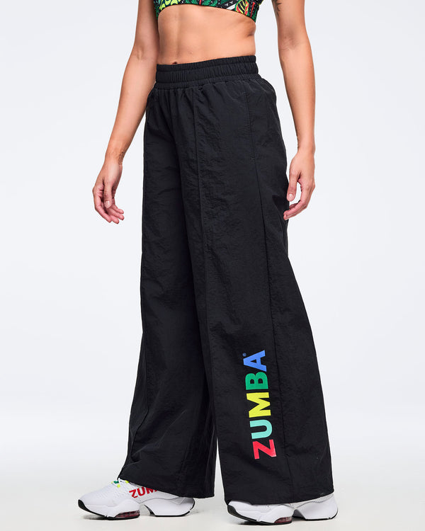 Black wide-leg Zumba pants with elastic waistband and multicolor vertical “ZUMBA” logo on the lower leg; female model pairs them with a tropical-print bra and white sneakers on a white background.