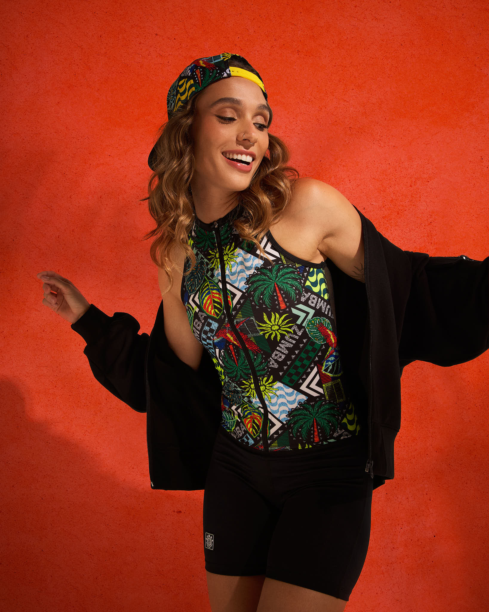 Sleeveless zip-front tropical-print bodysuit with black biker shorts, styled on a female wearing a black jacket and a printed cap against a textured orange backdrop.
