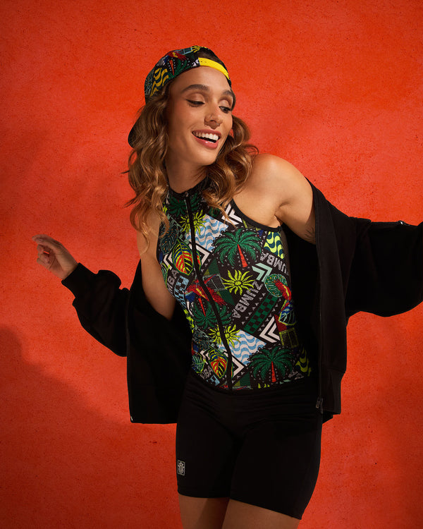 Sleeveless zip-front tropical-print bodysuit with black biker shorts, styled on a female wearing a black jacket and a printed cap against a textured orange backdrop.