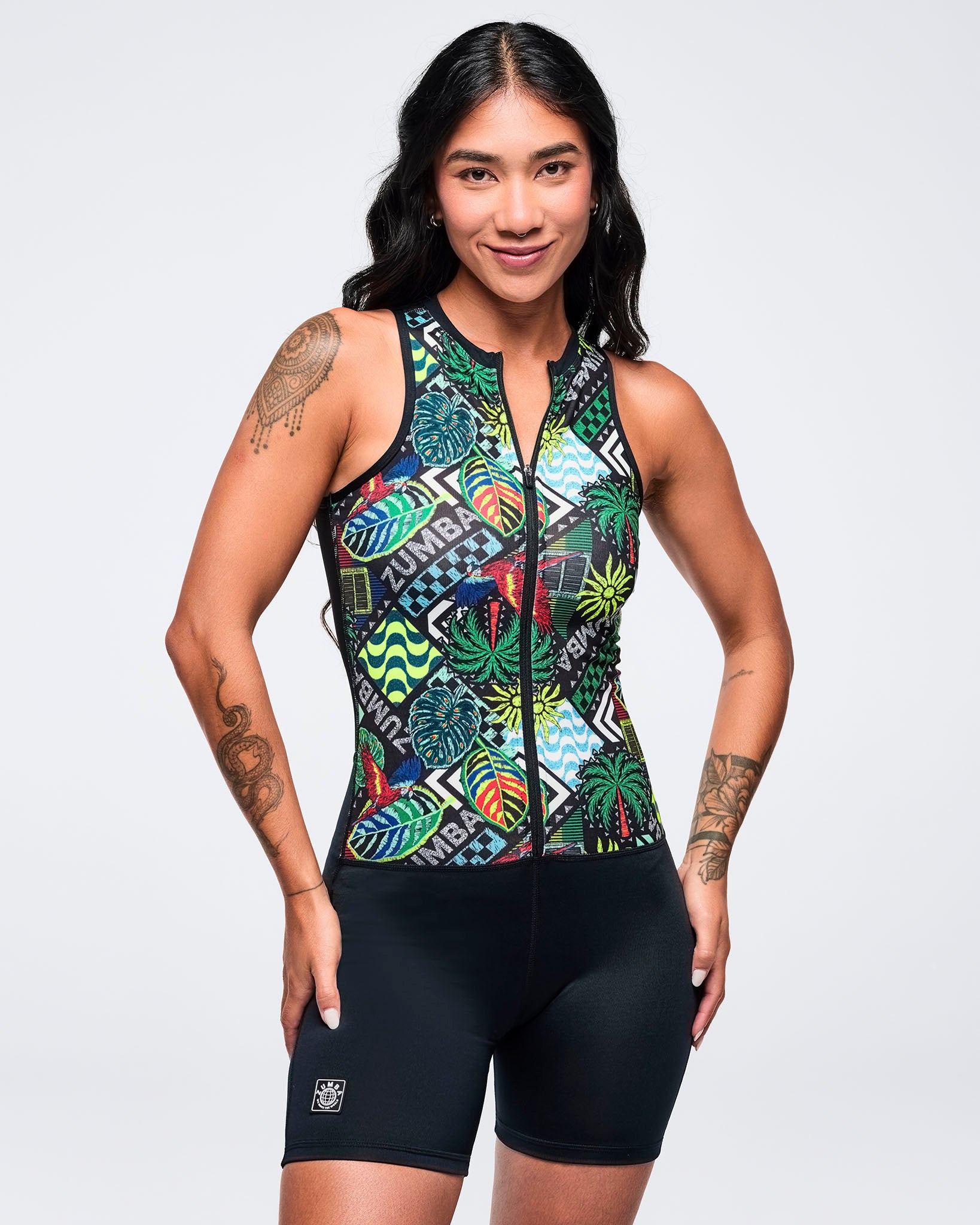 Sleeveless zip-front printed bodysuit with black biker-short legs and a colorful tropical collage pattern on the top, modeled by a female on a white studio background.