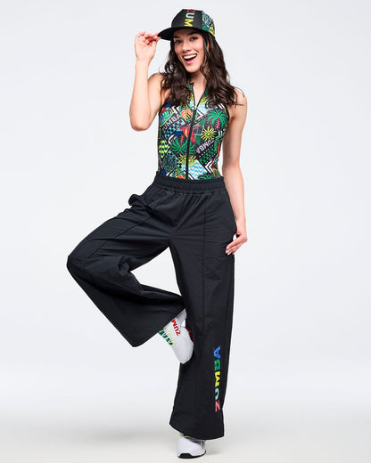Tropical-print sleeveless zip-front bodysuit, styled with black wide-leg pants featuring a multicolor “ZUMBA” leg graphic. Modeled by a female wearing a printed cap and white athletic sneakers on a white studio background.