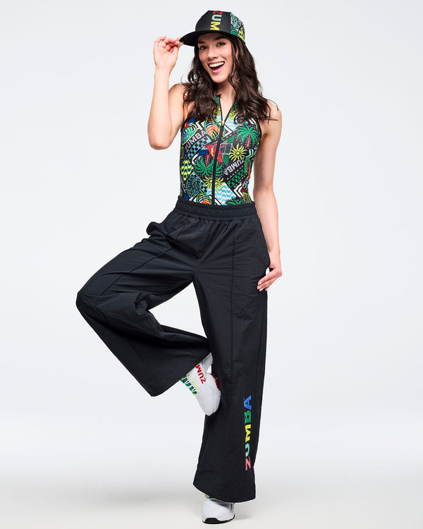 Tropical-print sleeveless zip-front bodysuit, styled with black wide-leg pants featuring a multicolor “ZUMBA” leg graphic. Modeled by a female wearing a printed cap and white athletic sneakers on a white studio background.