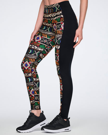 Side view of high-waisted full-length leggings with a multicolor geometric print on one side and a solid black panel on the other. Female model wears the fitted leggings with black athletic sneakers.