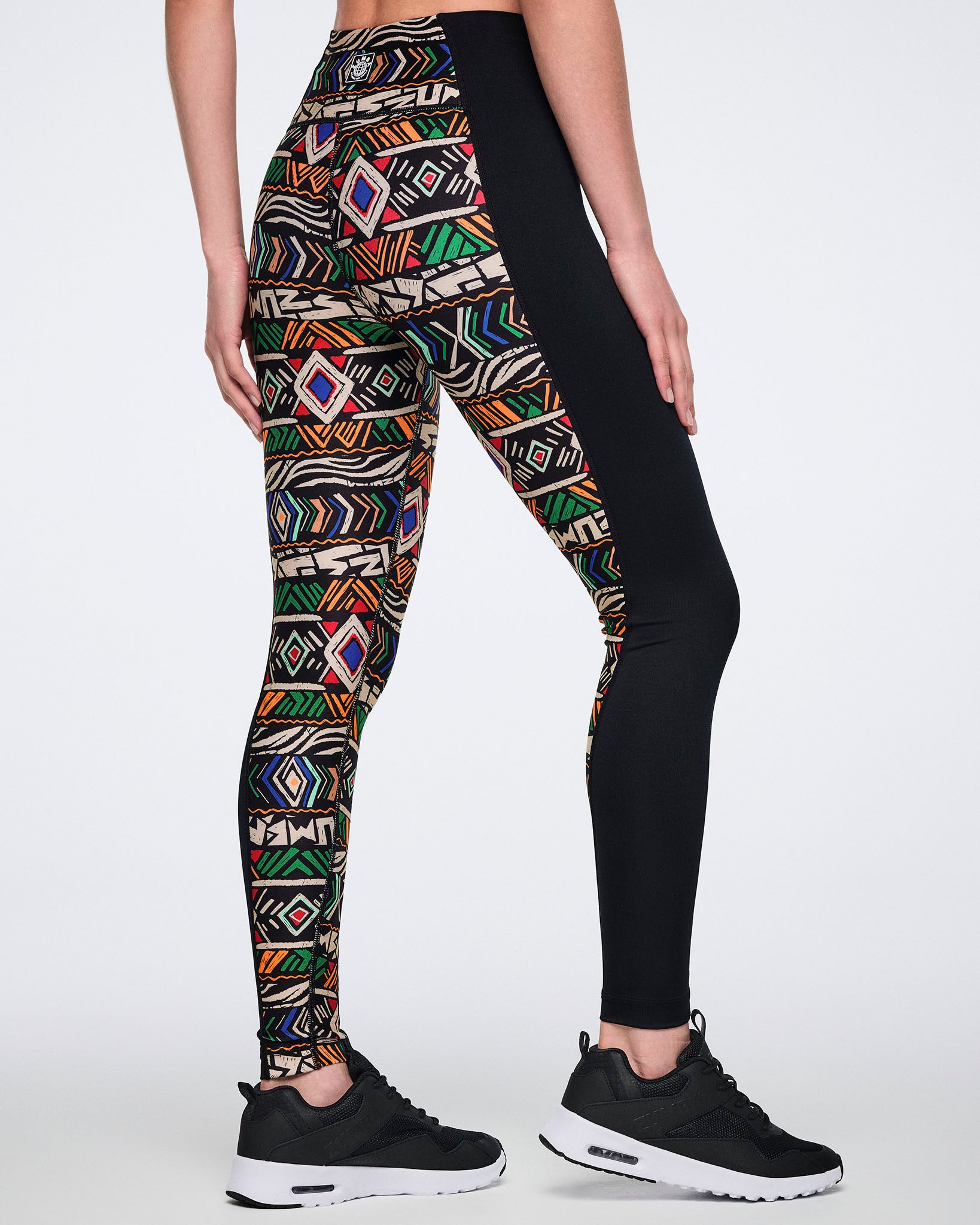 Back view of high-waisted leggings with a multicolor geometric print on one leg and solid black paneling on the other. Female model wears the fitted full-length leggings with black athletic sneakers.