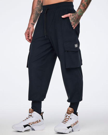 Black cargo jogger pants with oversized flap side pockets, a drawstring waistband, and ribbed ankle cuffs, shown on a male model wearing white high-top sneakers with black and leopard-print details.