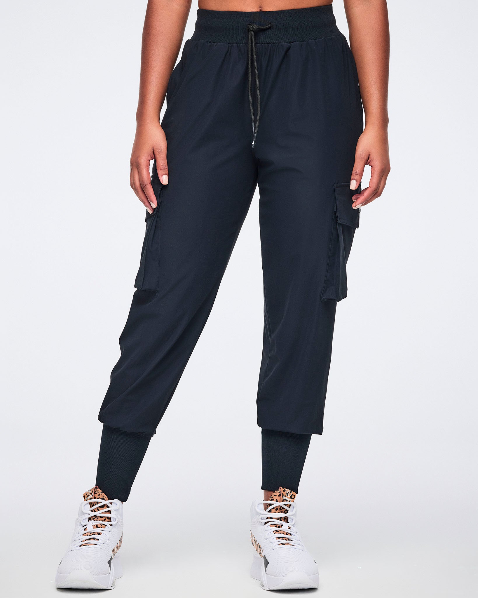 Black cargo jogger pants with oversized flap side pockets, a high-rise waistband with drawstring, and ribbed ankle cuffs, shown from the front on a female model wearing white high-top sneakers with black and leopard-print details.